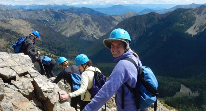 a group of students wearing helmets traverse a rocky landscape with mountains in the background